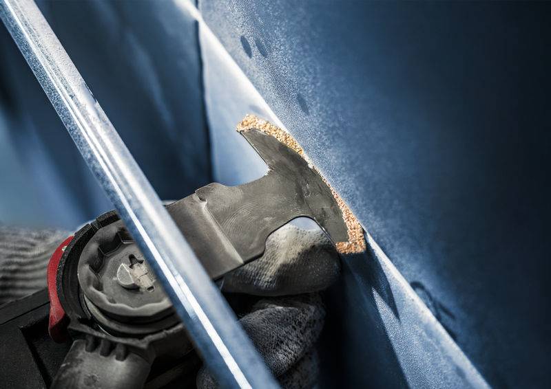 Eine Hand in einem Handschuh hält ein oszillierendes Werkzeug mit Schleifaufsatz, das eine blaue Oberfläche schleift.