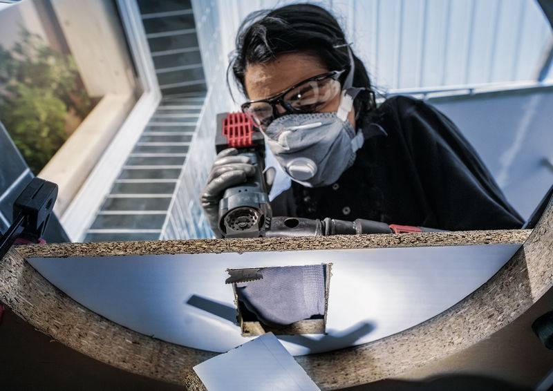 Eine Person trägt Schutzbrille und Atemmaske, während sie ein Loch in ein Stück Holz bohrt.