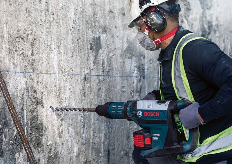 Ein Bauarbeiter mit Schutzhelm und Ohrenschützern bohrt mit einer Bosch-Bohrmaschine in eine Betonwand auf einer Baustelle.