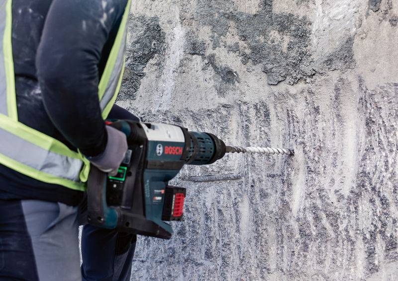 Eine Person bohrt mit einem Elektrowerkzeug in eine Betonwand. Die Bohrmaschine ist dunkelgrau-blau und wird mit beiden Händen gehalten.