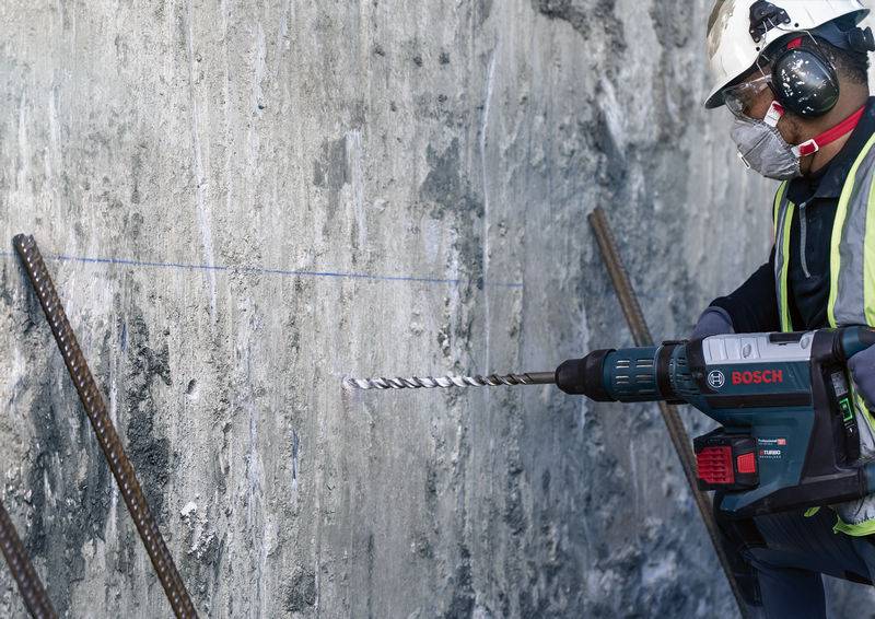 Arbeiter mit Schutzkleidung bohrt mit Bosch-Bohrer in Betonwand auf Baustelle. Betonstahl-Stäbe stehen aus der Wand heraus.