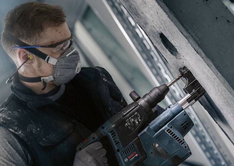 Ein Arbeiter trägt Schutzbrille und Maske, benutzt eine Elektrowerkzeug-Bohrmaschine, um ein Loch in eine Betonwand zu bohren.