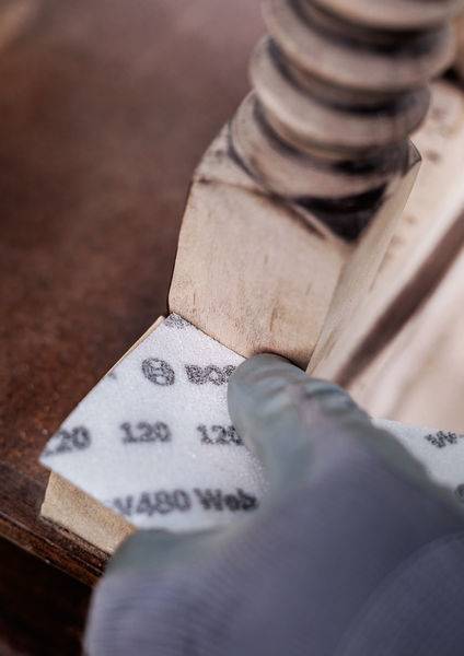 Eine Person schleift eine Holzoberfläche mit Schleifpapier. Das Schleifpapier ist zwischen einem dekorativen Möbelstück und einer Kante platziert.