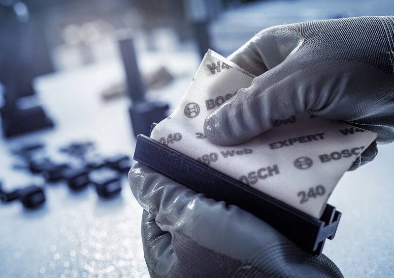 Eine Person trägt Handschuhe und poliert ein dunkles rechteckiges Objekt mit einem Schleifschwamm, auf dem 'Bosch Expert' steht.
