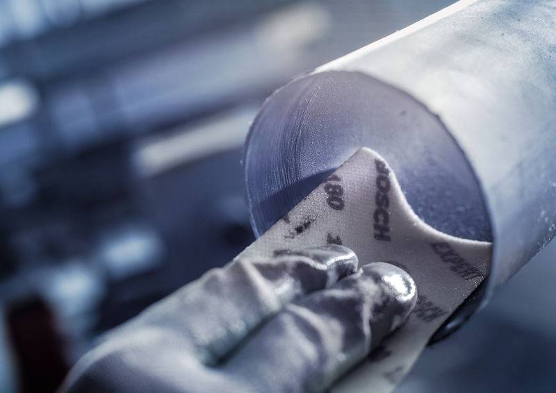 Eine Hand, die Schleifpapier auf einen großen Metallzylinder legt, zeigt eine Industrieszene mit Fokus auf Präzisionsarbeit.