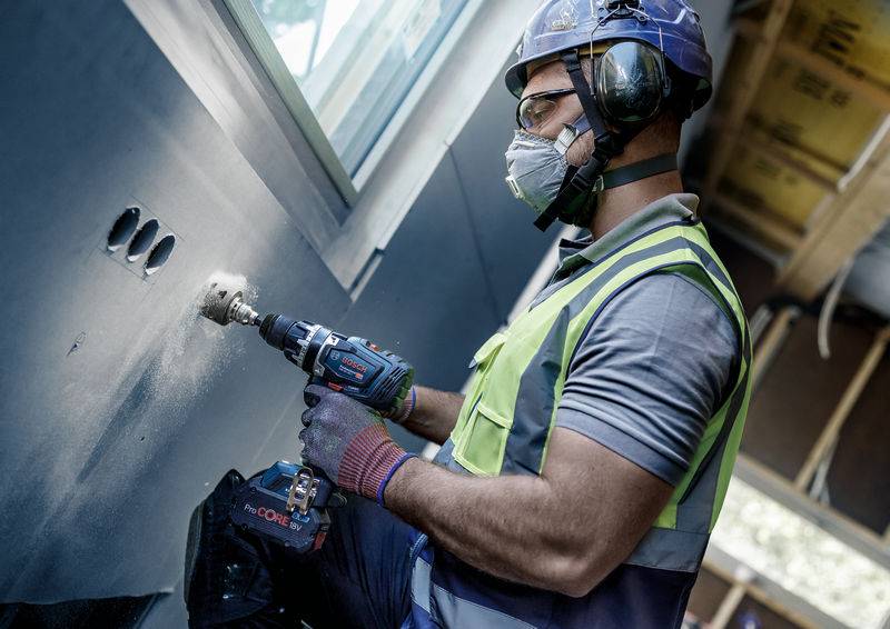 Ein Bauarbeiter in Schutzkleidung bohrt mit einem Elektrowerkzeug Löcher in eine Wand in einem hell beleuchteten Raum.