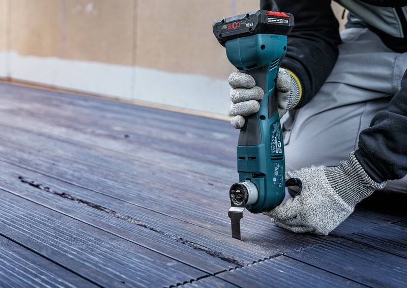 Eine Person arbeitet mit einem oszillierenden Multifunktionswerkzeug auf einer Holzterrasse, trägt Handschuhe und kniet.