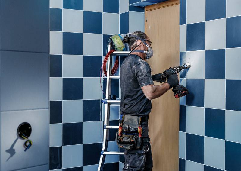 Eine Person in Arbeitskleidung bohrt mit einer Bohrmaschine ein Loch in eine türähnliche Fläche in einem Raum mit blau karierten Wänden.