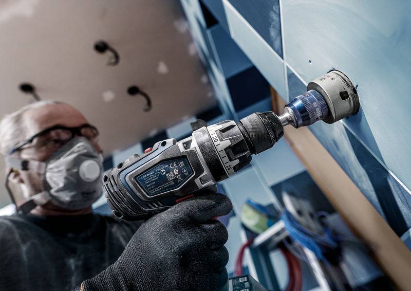Ein Handwerker bohrt mit einer elektrischen Bohrmaschine ein Loch in eine geflieste Wand. Er trägt eine Schutzmaske und Handschuhe.