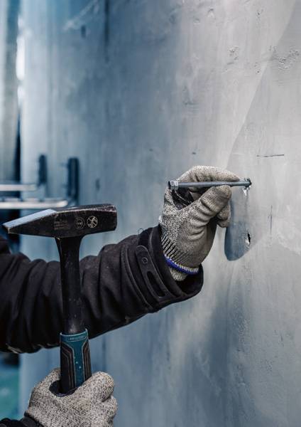 Eine Person trägt Handschuhe und hält einen Hammer, während sie einen Nagel in eine Wand schlägt.