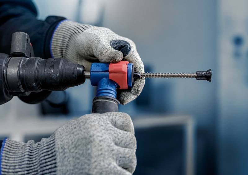 Eine Person hält ein Elektrowerkzeug mit Bohrer, trägt Arbeitshandschuhe und bohrt in ein Material. Hintergrund unscharf.