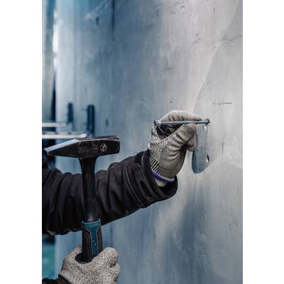 Eine Person schlägt mit einem Hammer einen Nagel in eine Betonwand, trägt dabei Handschuhe zum Schutz.
