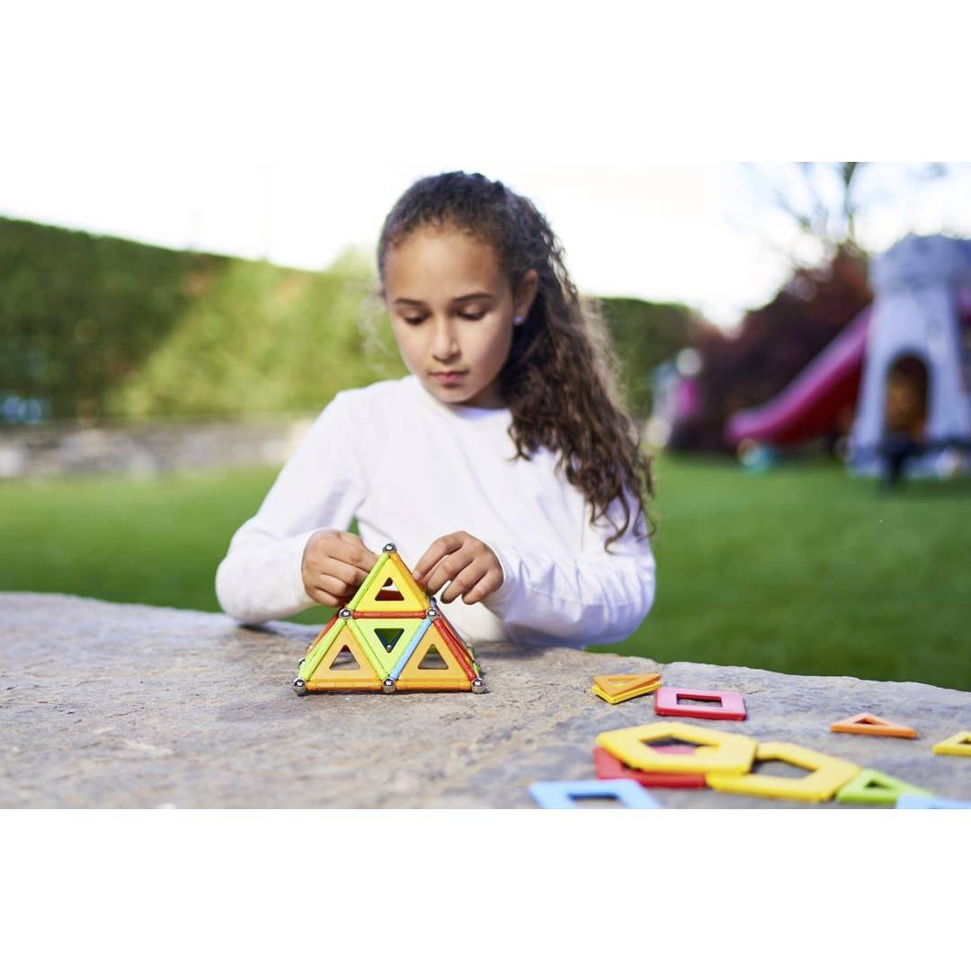 Mädchen spielt mit bunten geometrischen Magnetbausteinen auf einem Tisch im Freien. Hintergrund unscharf mit Rasen und Rutsche.