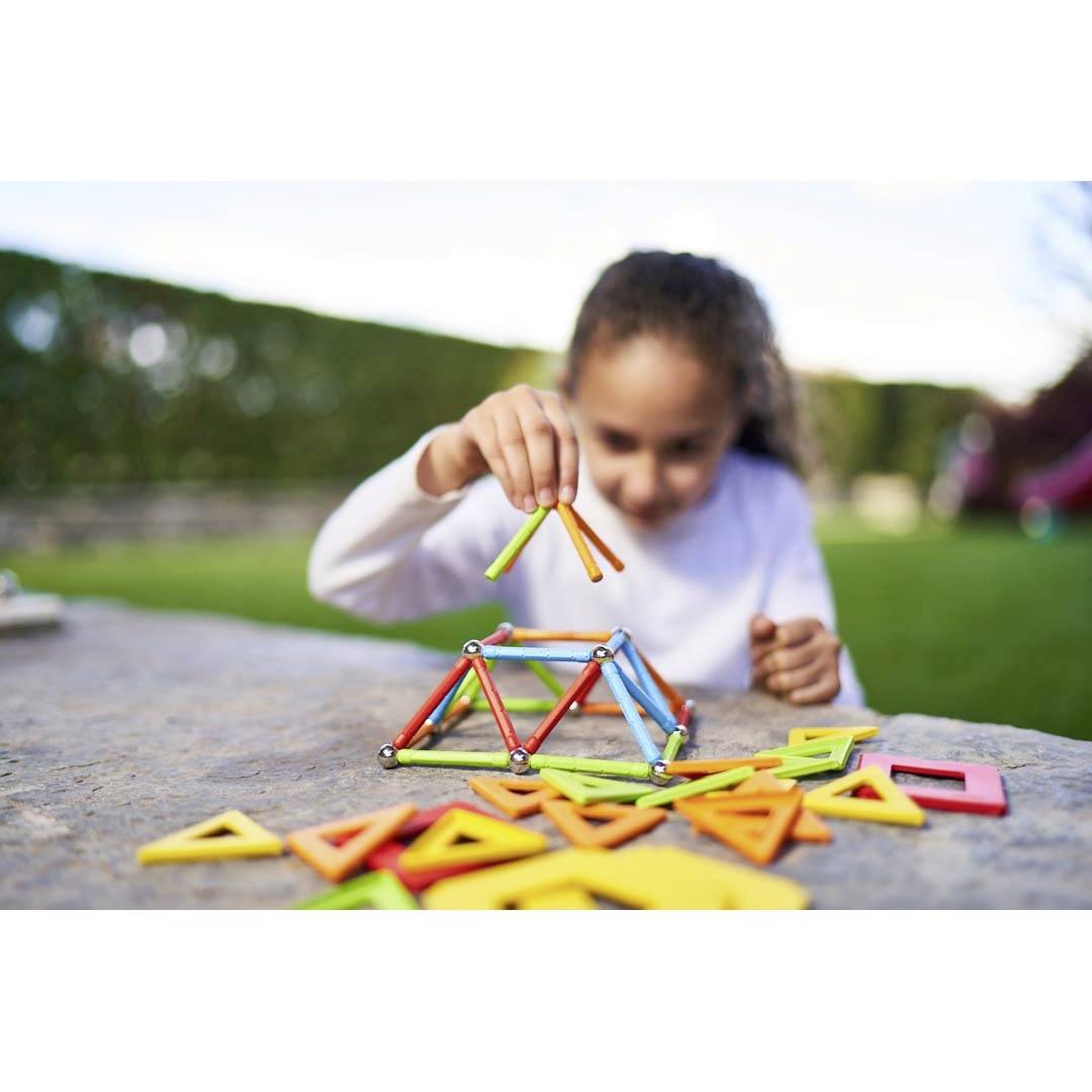 Ein Kind spielt im Freien mit bunten magnetischen Bausteinen und baut ein kleines Modell. Der Hintergrund ist ein grüner Garten.