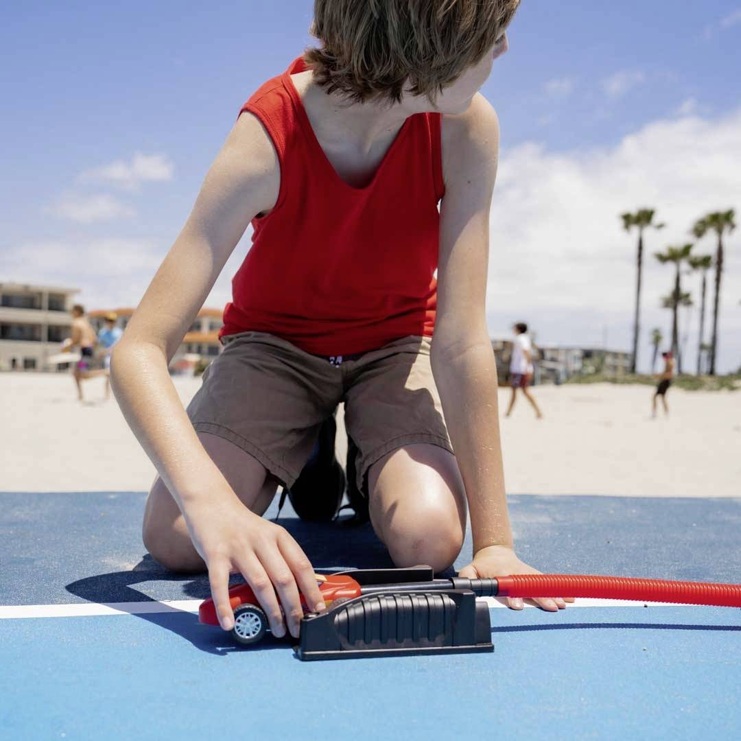 Ein Kind in rotem T-Shirt und Shorts spielt mit einer roten Pumpe auf einem blauen Boden am Strand. Im Hintergrund Palmen und Gebäude.