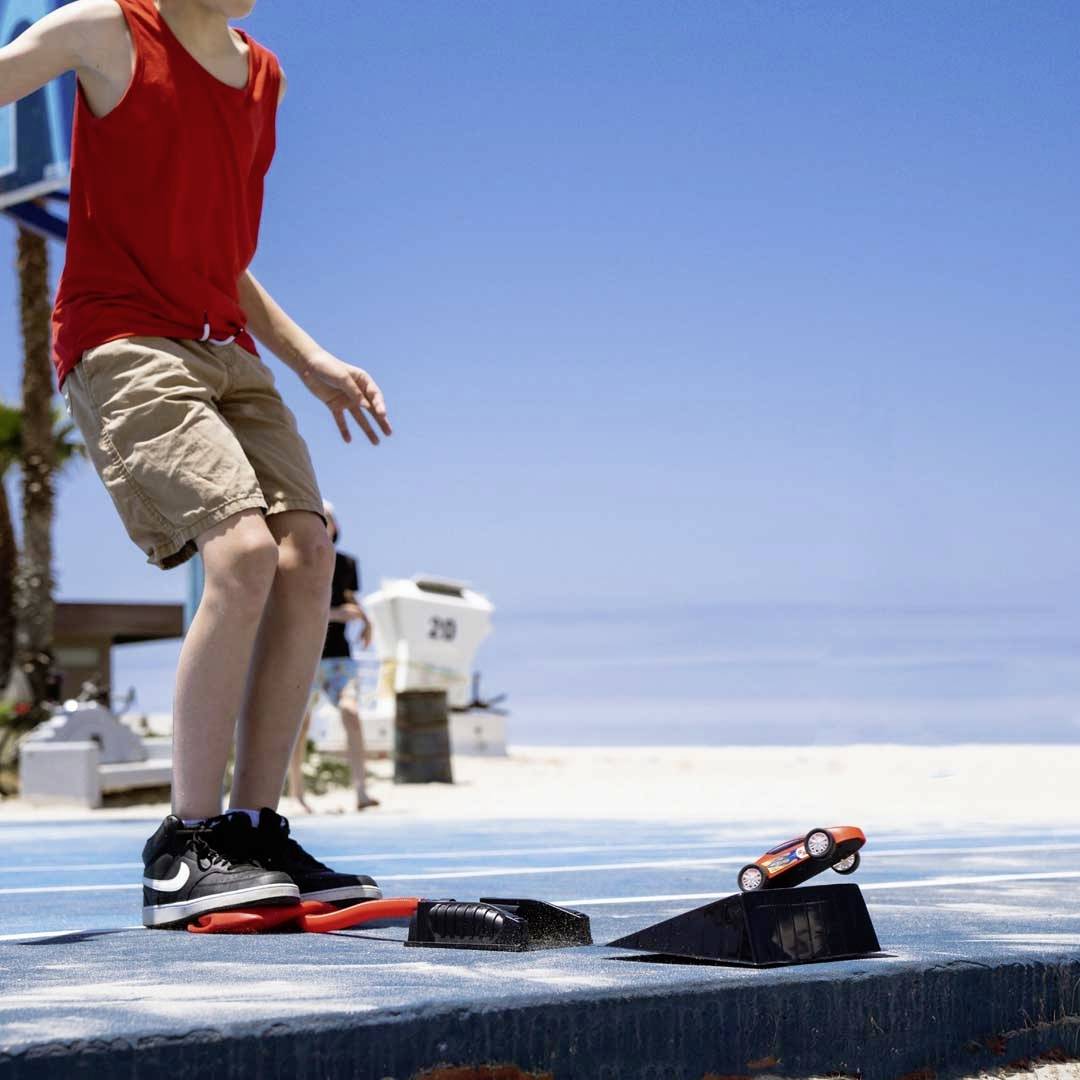 Ein Kind mit rotem Oberteil und beigen Shorts fährt auf einem Skateboardrampe am Strand. Im Hintergrund sind Palmen zu sehen.