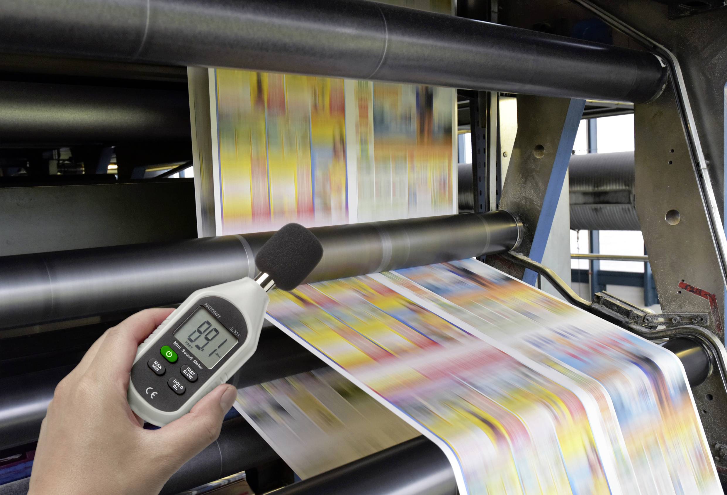 Druckmaschine in Aktion mit Fokus auf einer Hand, die ein Dezibel-Messgerät hält, das 89,1 Dezibel anzeigt.