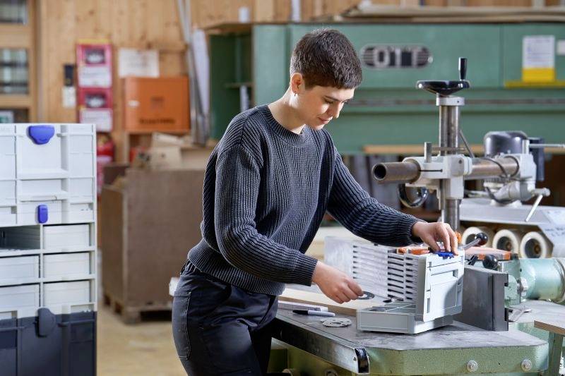 Eine Person in einer Werkstatt, die einen dunklen Pullover trägt, an einem Tisch mit einer Maschine arbeitet, umgeben von Werkzeugen und Lagerboxen.