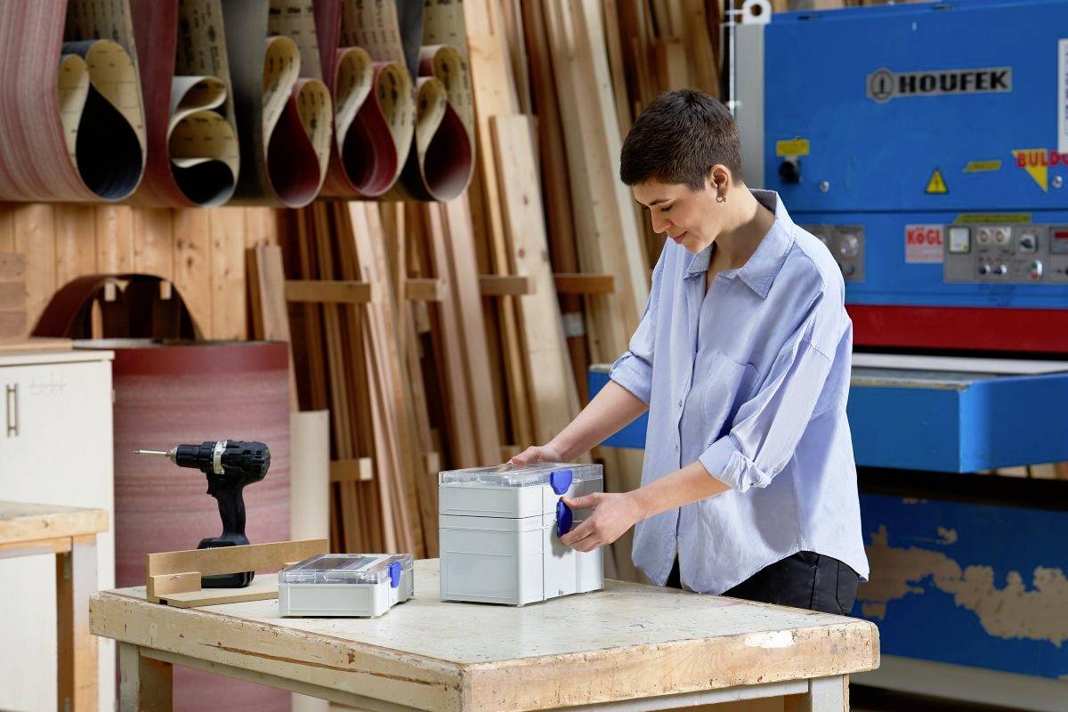 Eine Person in einer Werkstatt schleift eine Holzkiste mit einem blauen Werkzeug, umgeben von Holzbearbeitungsgeräten und Furnierschichten.