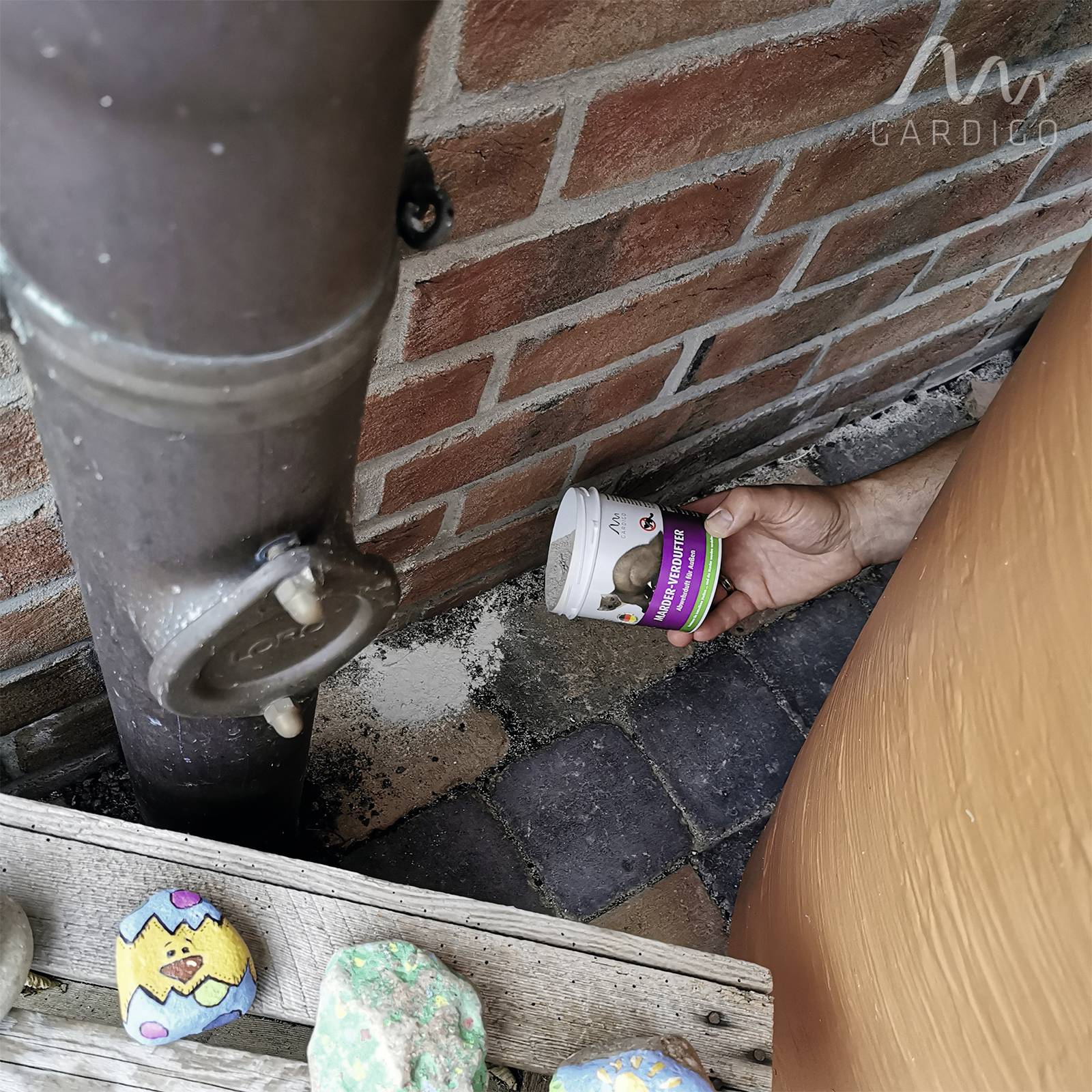 Eine Hand greift hinter ein Regenrohr nach einem Plastikbecher mit Gesichtsmotiv, nahe einer Backsteinwand. Im Vordergrund bemalter Stein.