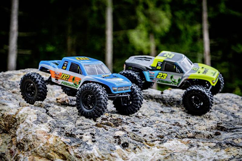 Zwei ferngesteuerte Modellautos fahren über felsiges Gelände im Wald. Ein Auto ist blau, das andere grün. Beide haben große Reifen.