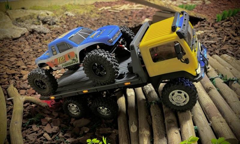 Ein ferngesteuerter gelber LKW auf einem hölzernen Weg transportiert ein kleineres, blaues Auto über unwegsames Gelände.