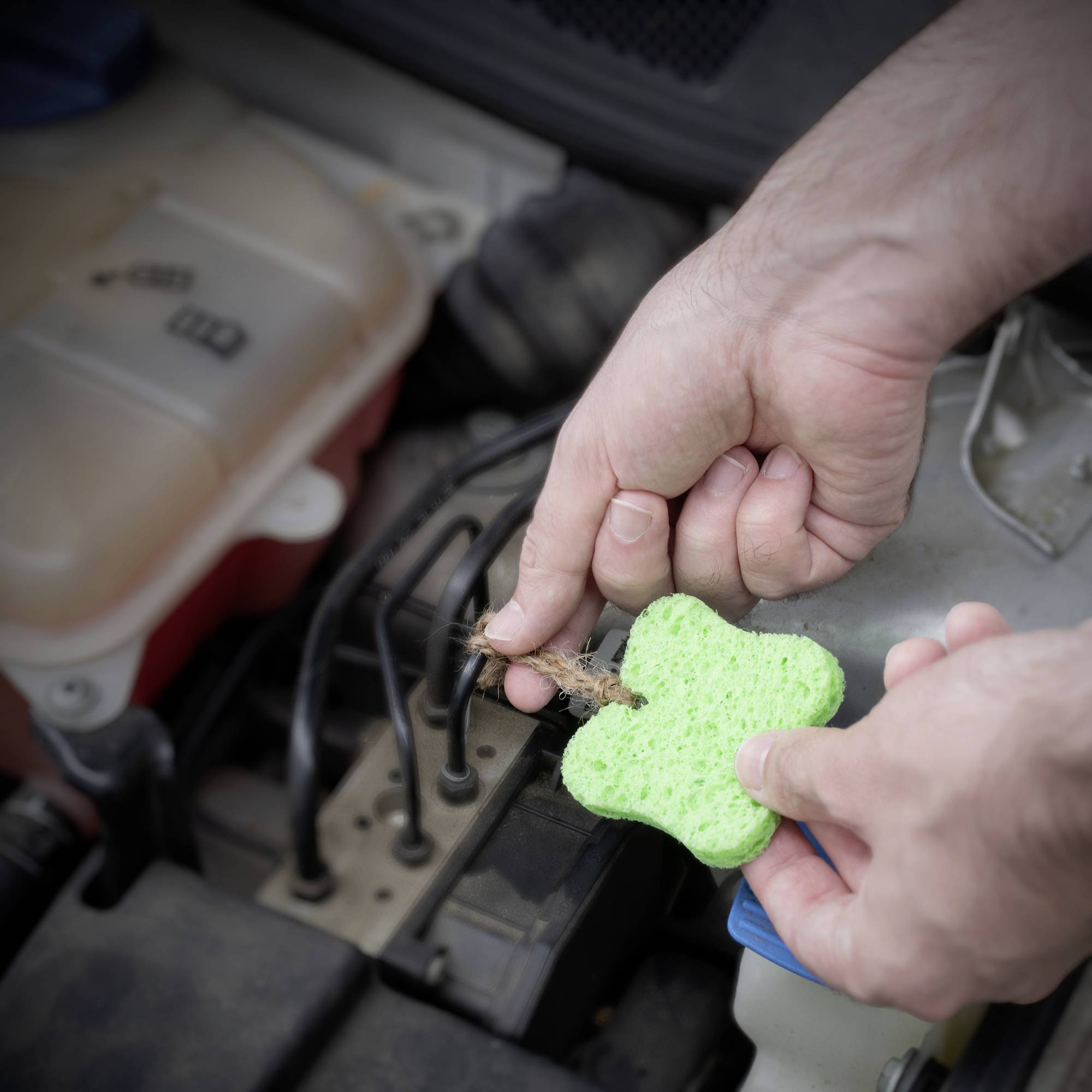 Eine Person hält einen grünen Schwamm über einem geöffneten Autoteilmotor, möglicherweise um ihn zu reinigen oder zu warten.