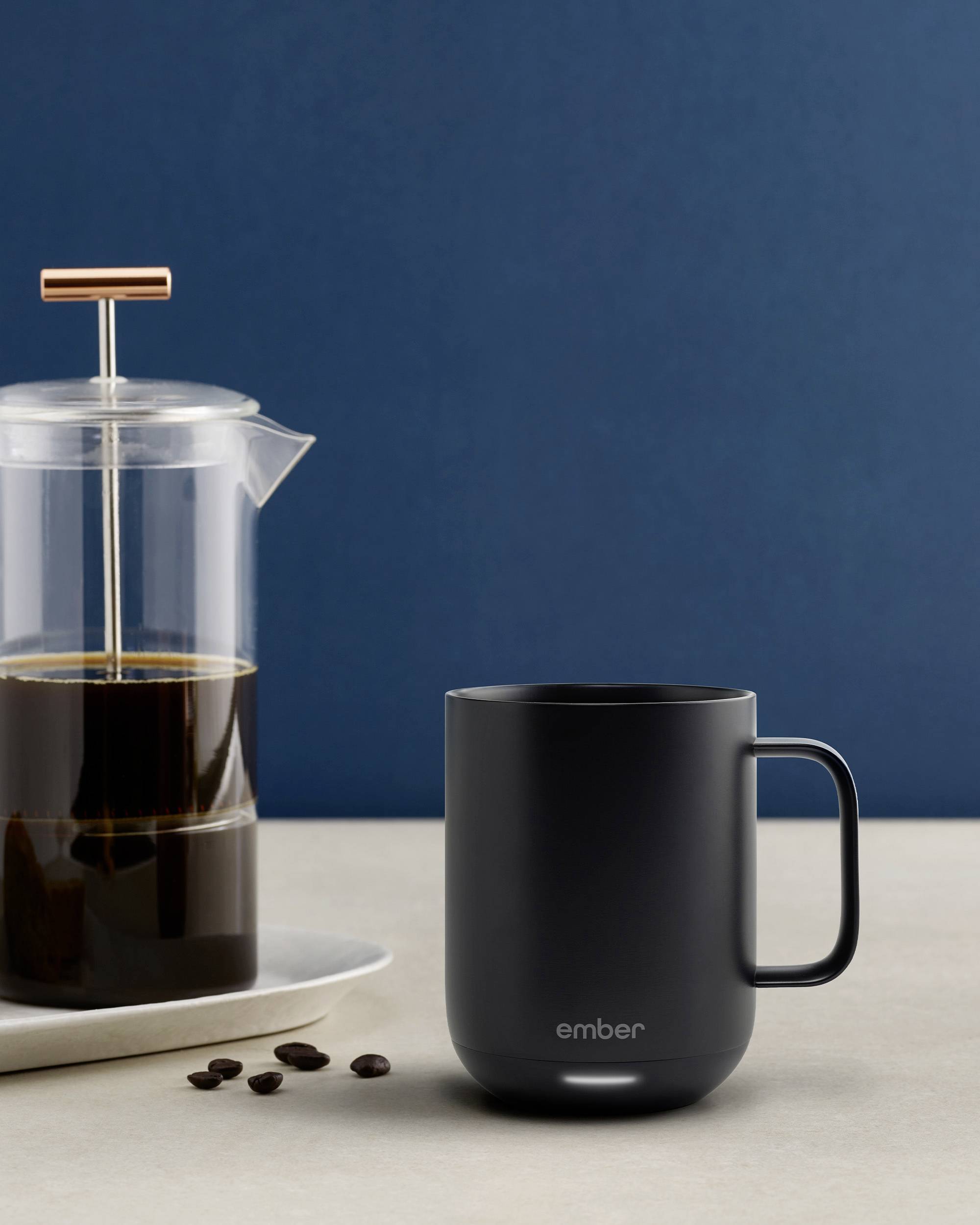 Schwarze Kaffeetasse auf hellem Tisch, neben einer transparenten French Press mit schwarzem Kaffee. Hintergrund in dunklem Blau.