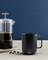 Schwarze Kaffeetasse auf hellem Tisch, neben einer transparenten French Press mit schwarzem Kaffee. Hintergrund in dunklem Blau.
