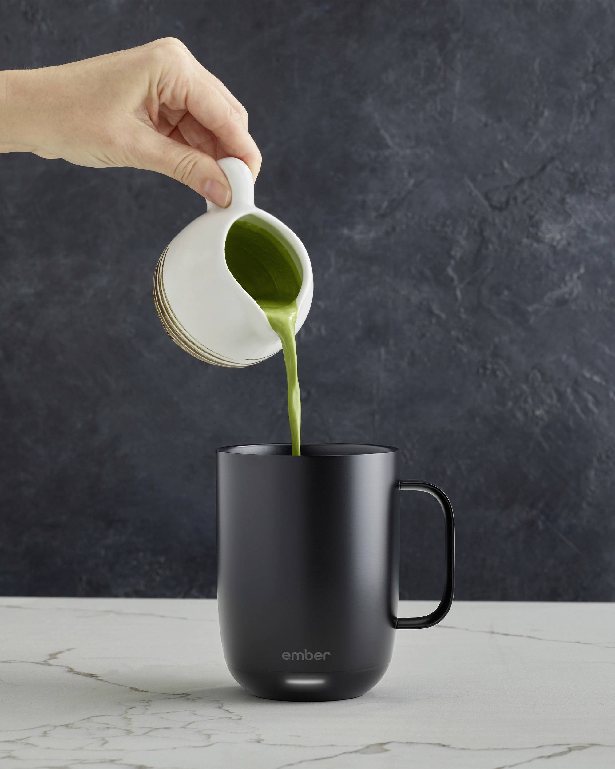 Eine Hand gießt Matcha aus einem weißen Krug in eine schwarze Tasse auf einem Marmortisch.