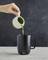 Eine Hand gießt Matcha aus einem weißen Krug in eine schwarze Tasse auf einem Marmortisch.