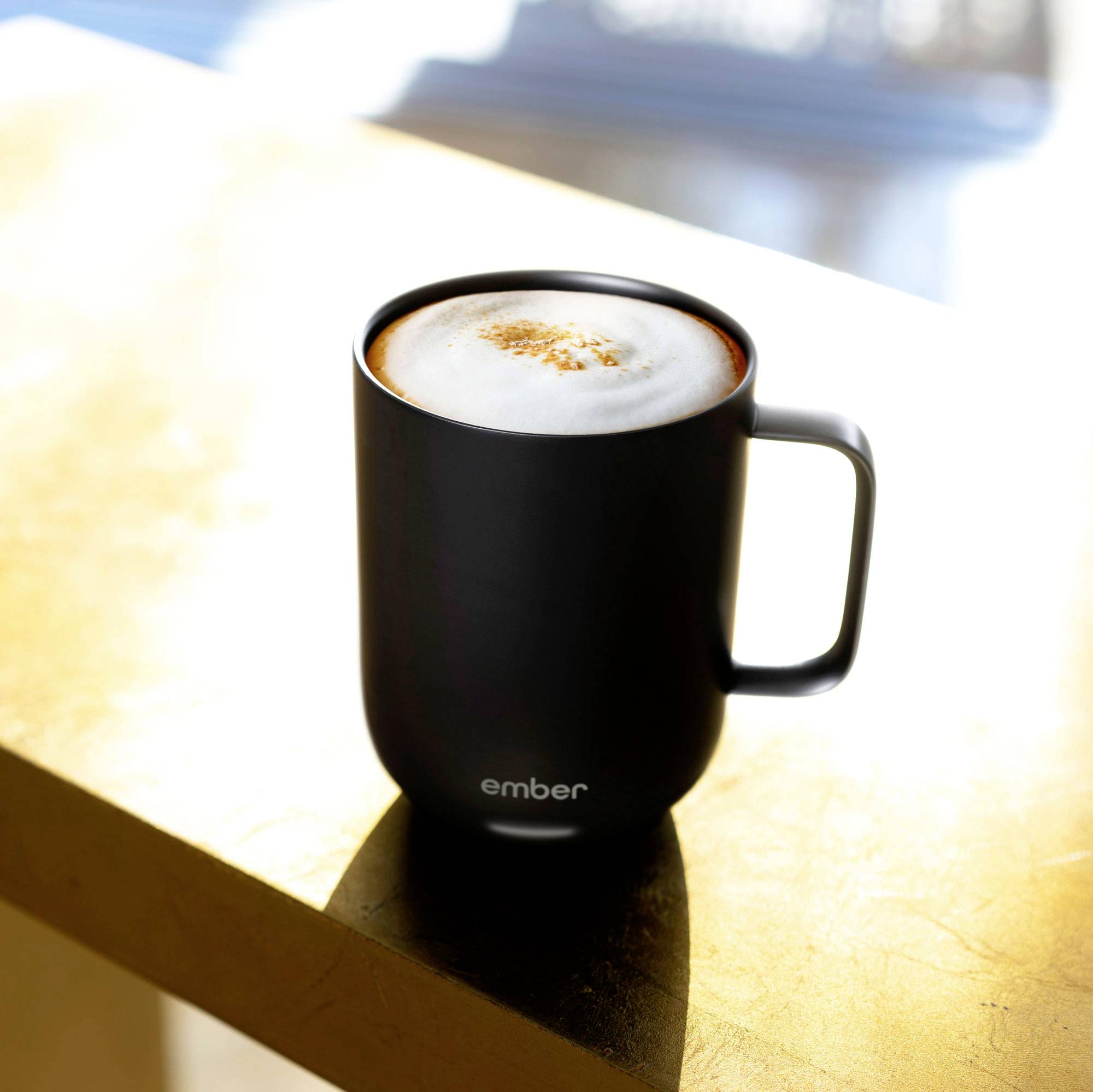 Eine schwarze Tasse mit 'ember'-Logo, gefüllt mit Schaumkaffee, steht auf einem Tisch in sonnigem Licht.