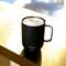 Eine schwarze Tasse mit 'ember'-Logo, gefüllt mit Schaumkaffee, steht auf einem Tisch in sonnigem Licht.