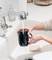Eine Person wäscht eine schwarze Tasse unter fließendem Wasser in der Spüle. Im Hintergrund sind Seifenspender und Geschirr sichtbar.
