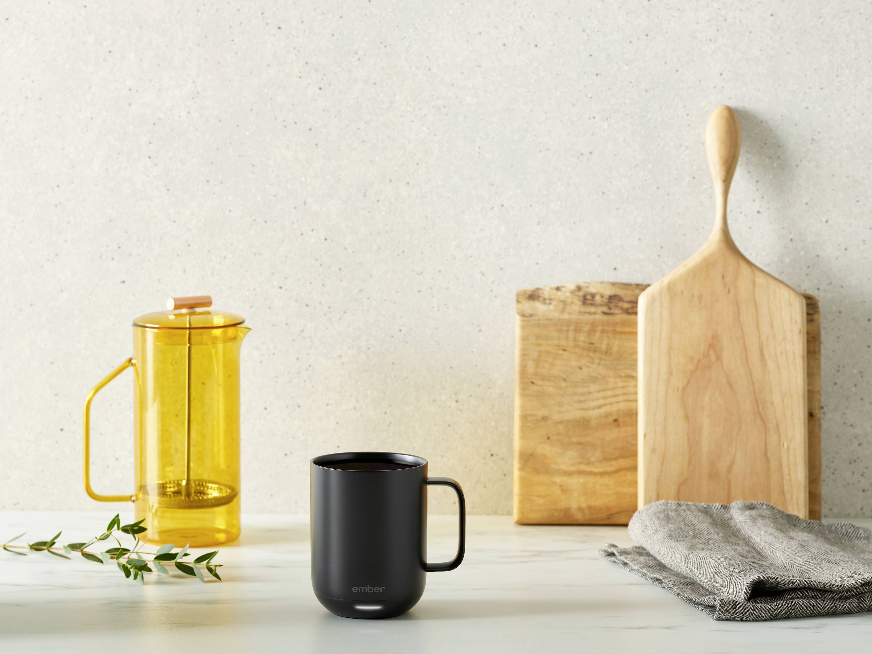 Eine schwarze Tasse steht auf einem Tisch neben einer gelben Kaffeepresse, einem Holzschneidebrett und einem grauen Handtuch.