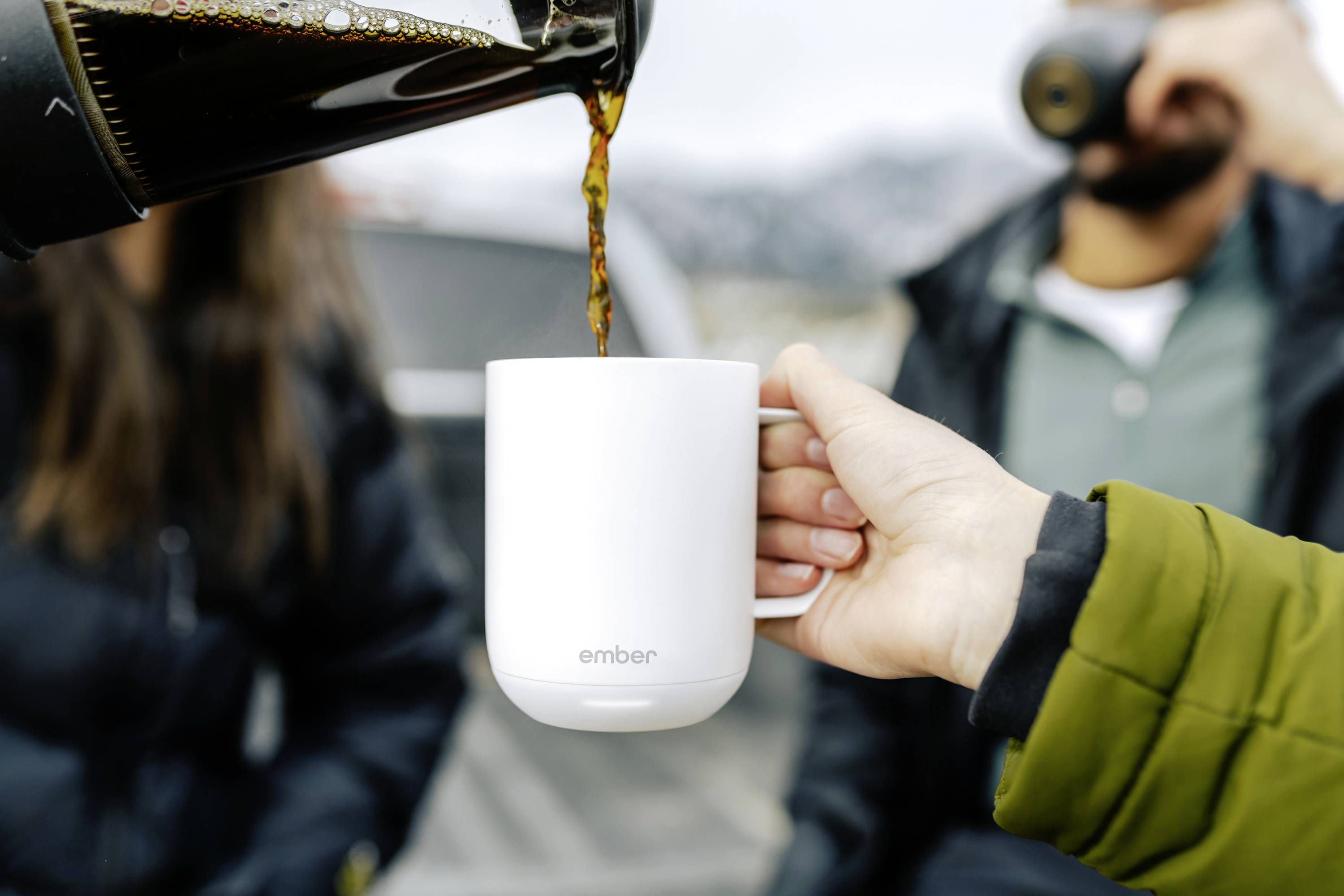 Eine Hand hält eine weiße Tasse mit der Aufschrift 'ember', in die Kaffee aus einer Kanne eingeschenkt wird. Im Hintergrund sind verschwommen Personen zu sehen.