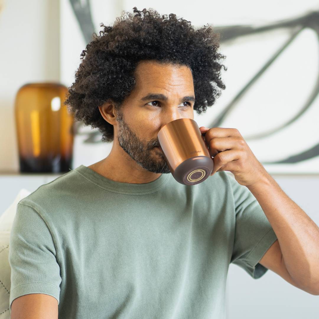 Eine Person mit lockigem Haar trinkt aus einer bronzefarbenen Tasse. Im Hintergrund sind verschwommen eine Vase und Kunstwerke zu sehen.
