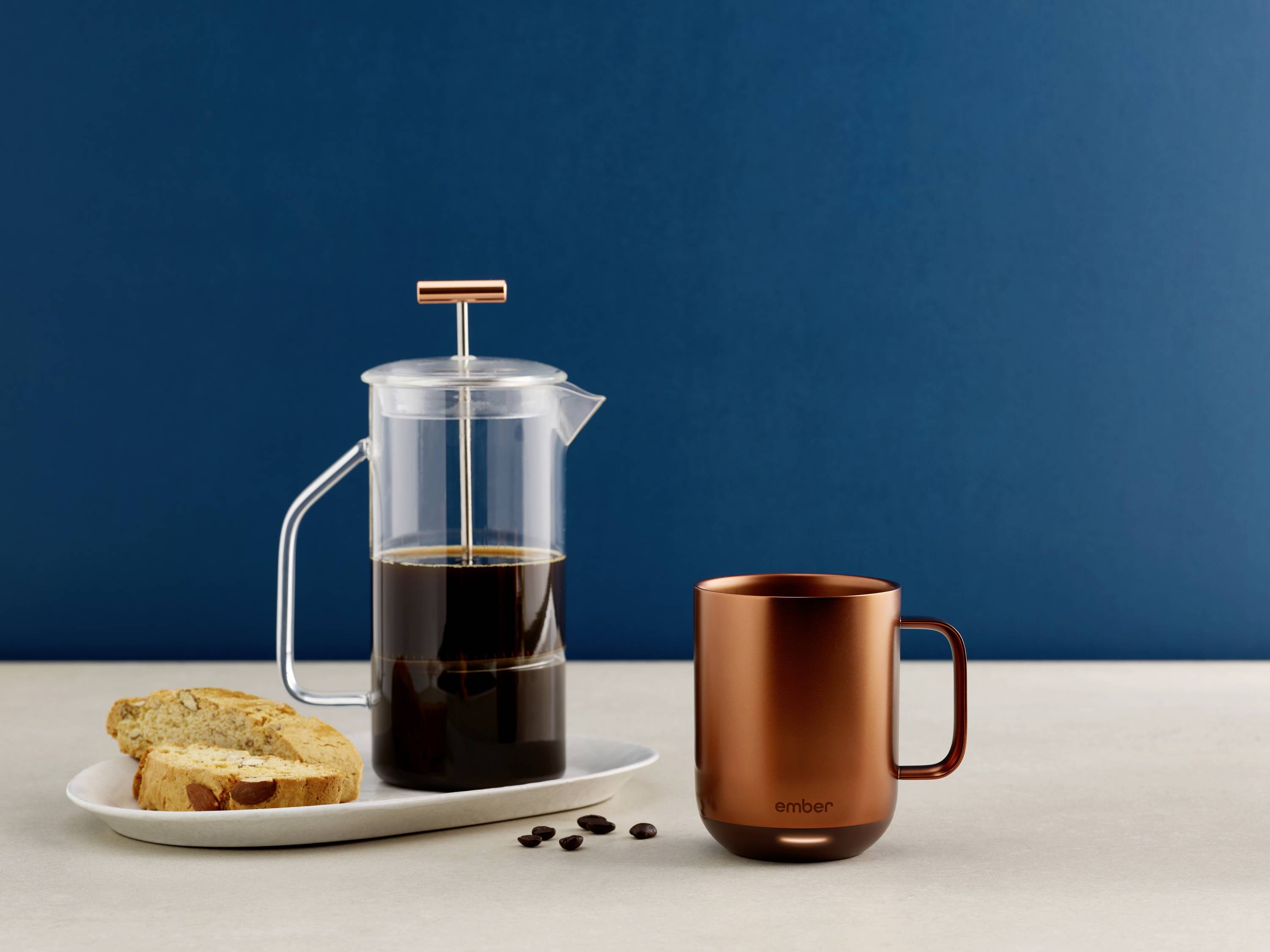 Eine Glaskanne mit Kaffee und ein kupferfarbener Becher stehen auf einem Tisch. Daneben liegt ein Teller mit zwei Scheiben Brot.