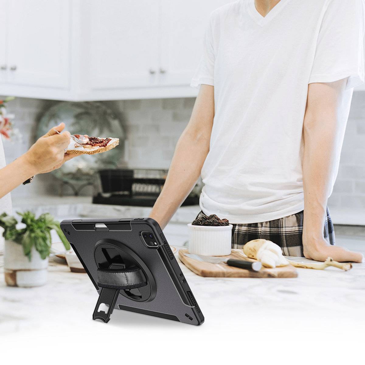 Eine Person hält ein Tablett mit einer Schutzhülle in einer modernen Küche, während zwei Personen im Hintergrund auf einer Marmor-Arbeitsplatte Essen zubereiten.