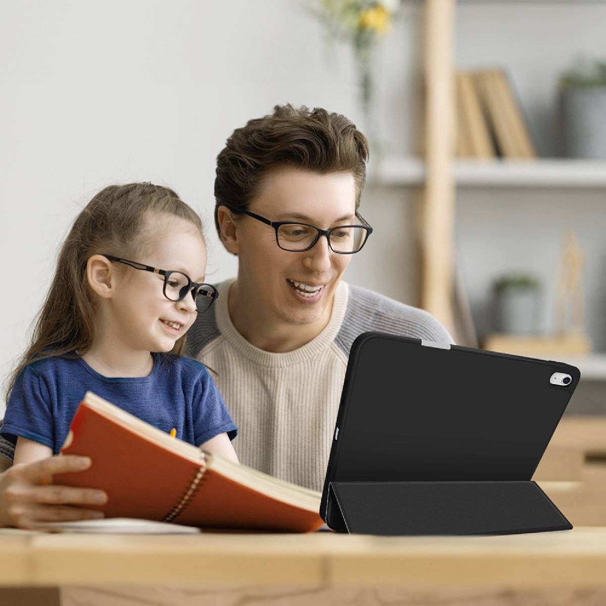 Ein Erwachsener und ein Kind sitzen am Tisch, lachen und schauen auf ein Tablet, während das Kind ein Buch hält.