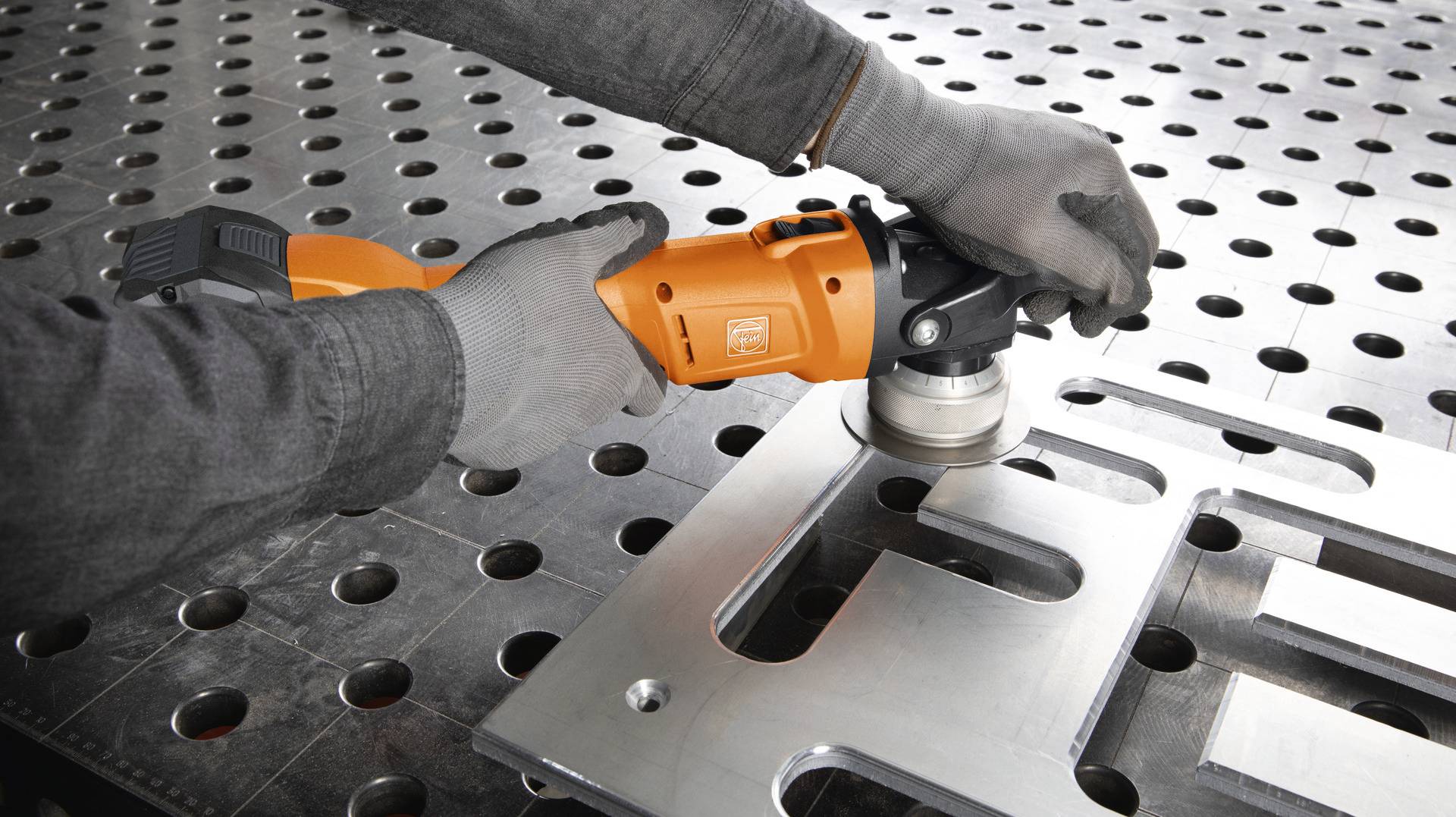Eine Person verwendet einen Winkelschleifer, um ein Metallteil auf einem Arbeitstisch mit Lochmuster zu bearbeiten.