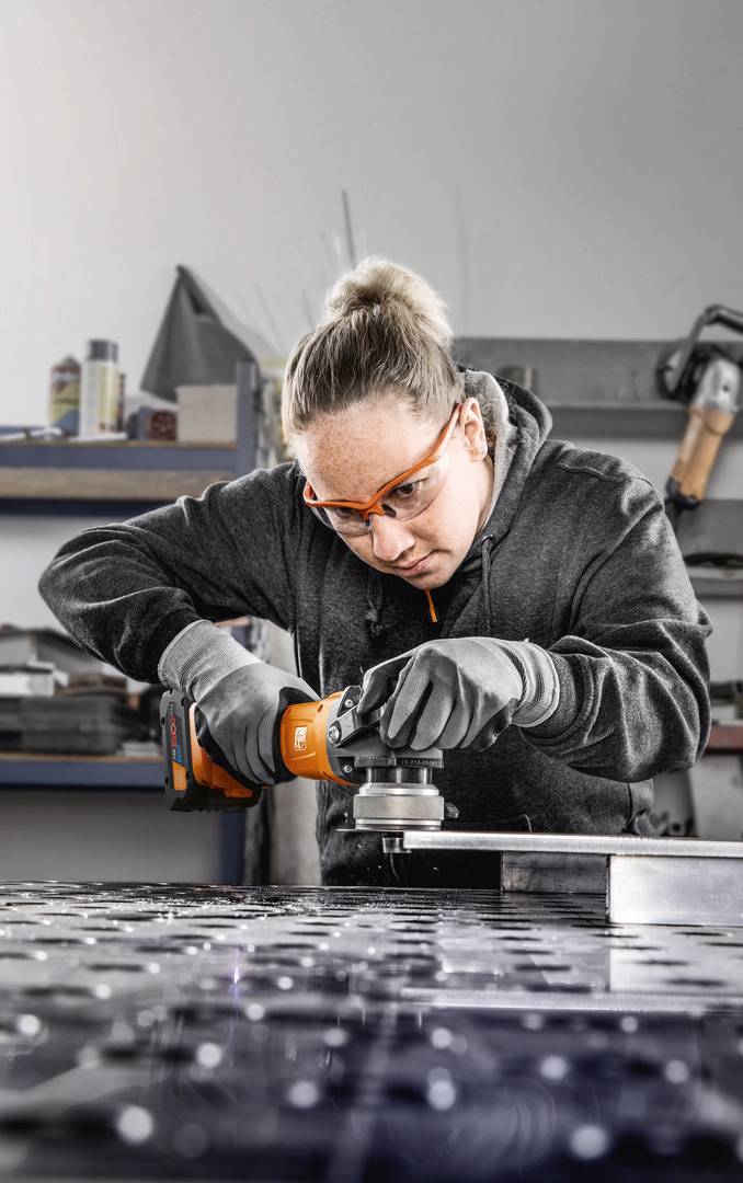 Eine Person trägt Schutzbrille und verwendet ein Schleifwerkzeug, um Metall auf einer Werkbank in einer Werkstatt zu bearbeiten.