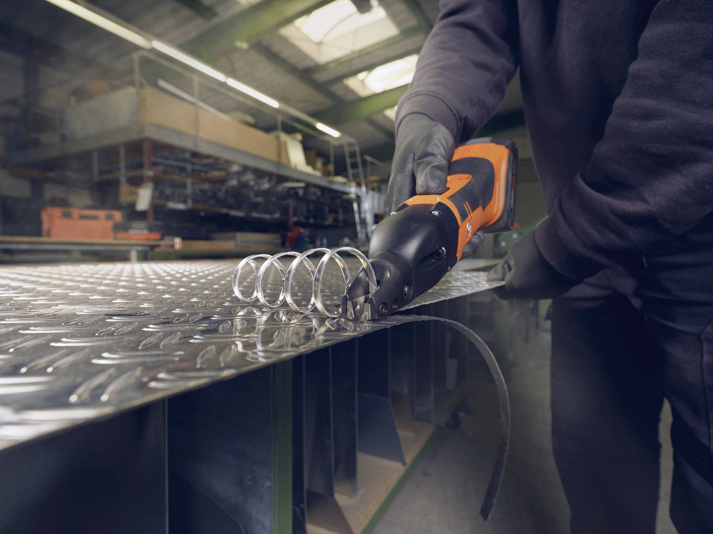 Eine Person schneidet mit einer Akku-Blechschere in einer Werkstatt ein Metallblech. Im Hintergrund sind Regale und Werkzeuge zu sehen.