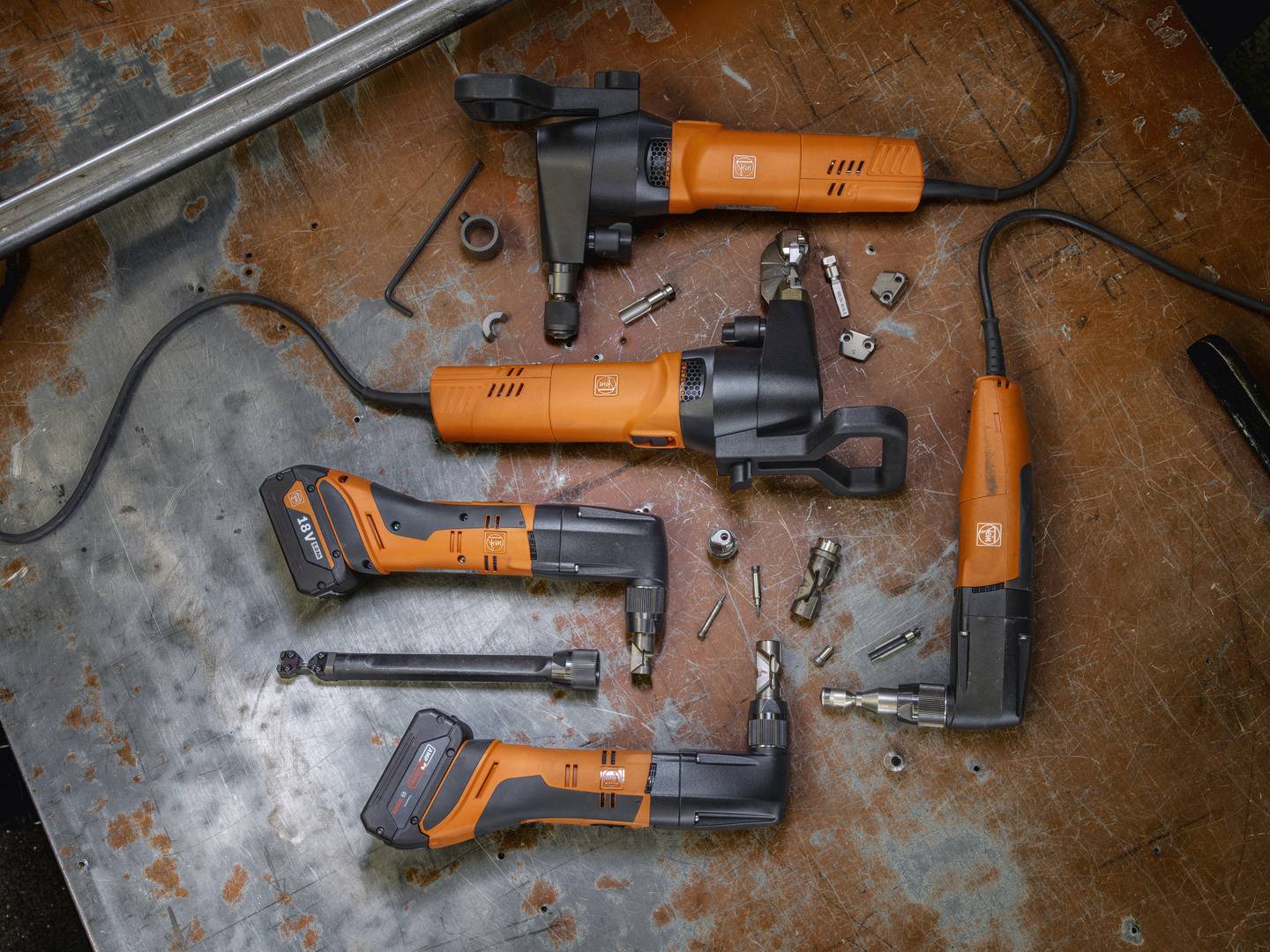 Mehrere orangefarbene Elektrowerkzeuge liegen auf einer Metalloberfläche, umgeben von Schrauben und Muttern. Werkstattumgebung.