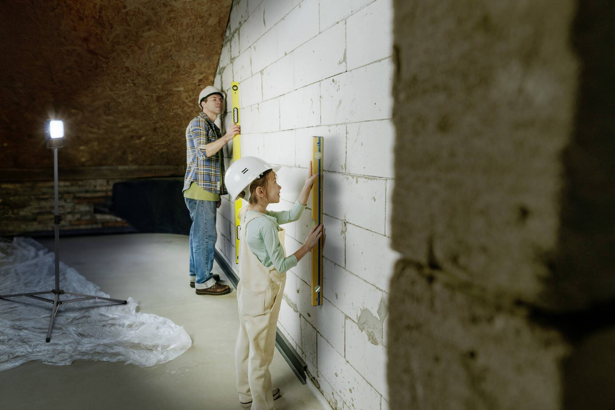 Zwei Personen mit Schutzhelmen messen eine Wand mit Wasserwaagen in einem Raum mit Betonwänden und einer Lampe zur Beleuchtung.