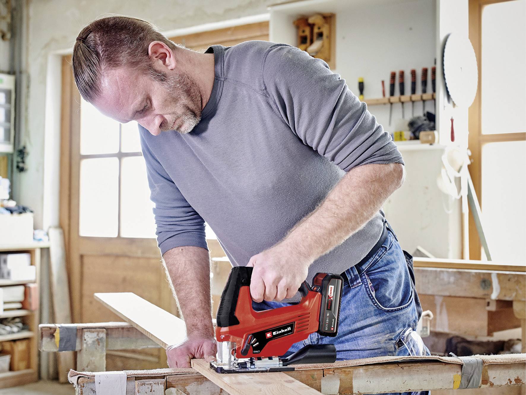 Ein Mann arbeitet in einer Werkstatt mit einer roten elektrischen Säge an einem Holzbrett. Im Hintergrund sind Werkzeuge an der Wand.