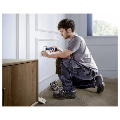 Ein Mann repariert eine Steckdose an einer Wand. Er kniet auf dem Boden, hält ein Werkzeug in der Hand, neben ihm liegen weitere Werkzeuge.