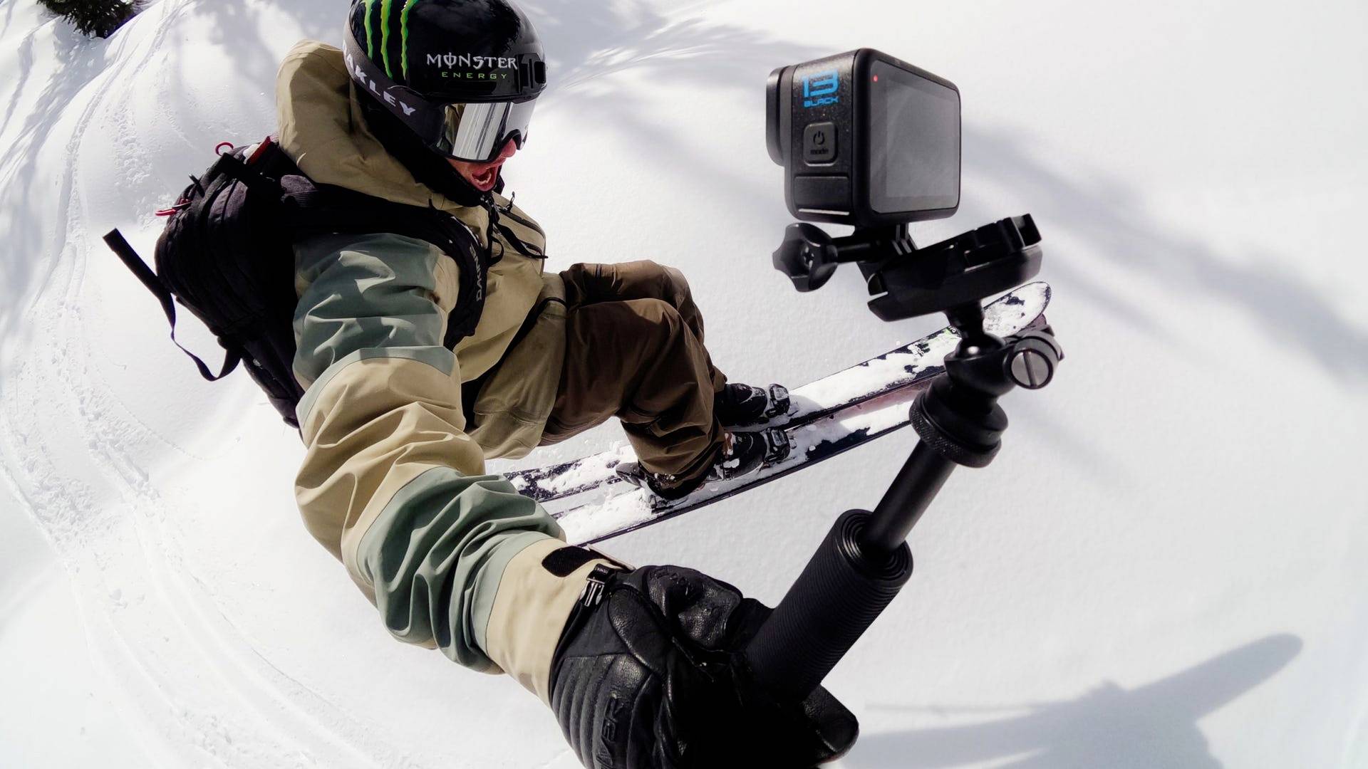 Ein Snowboarder fährt durch frischen Pulverschnee, hält eine Action-Kamera auf einem Selfie-Stick, um die Fahrt zu filmen.