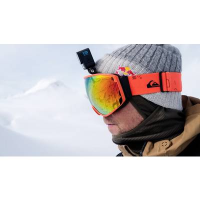 Eine Person in Winterkleidung sieht nach rechts. Sie trägt eine Orange Ski-Brille und eine graue Mütze mit einer Kamera darauf. Schneebedeckte Berge im Hintergrund.