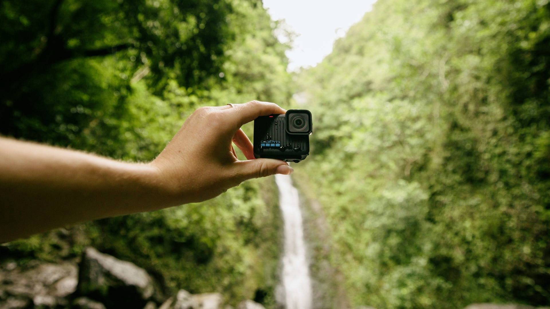 Eine Hand hält eine Actionkamera vor einem unscharfen Hintergrund mit einem Wasserfall und dichtem Wald.